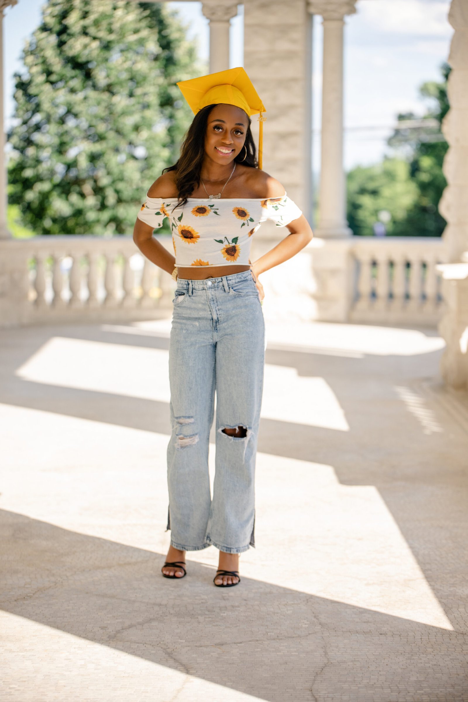 Graduate in a gold cap and sunflower top smiling under a sunlit colonnade during her graduation portrait session