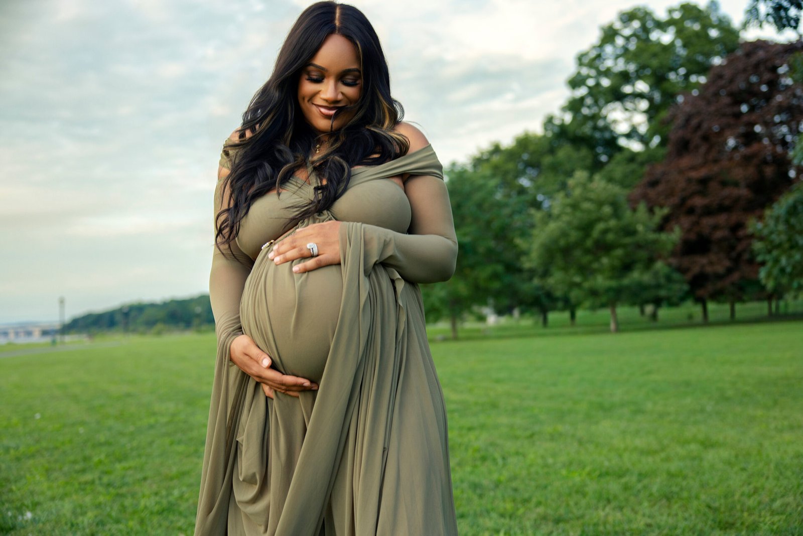 Smiling mother-to-be in a flowing olive gown cradling her belly in a riverside park at the Mother’s Blessing