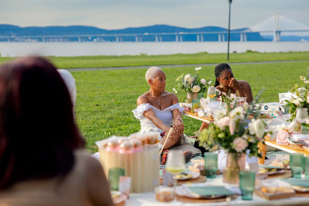 Guests listening at a riverside Mother’s Blessing picnic hosted by The Christian Doula Experience, with the Hudson River in the background