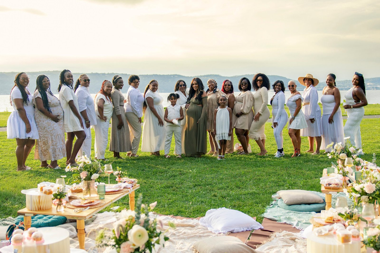 Group photo of guests in white and neutral outfits surrounding the mother-to-be at a riverside Mother’s Blessing picnic hosted by The Christian Doula Experience