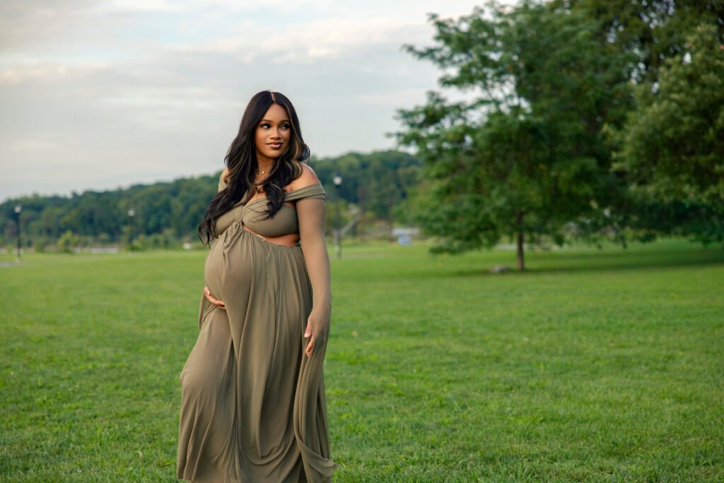Mother-to-be in a flowing olive gown walking through a riverside park and glancing over her shoulder at the Mother’s Blessing