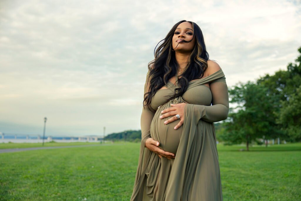 Mother-to-be in an olive gown cradling her belly beneath an open sky in a riverside park at the Mother’s Blessing