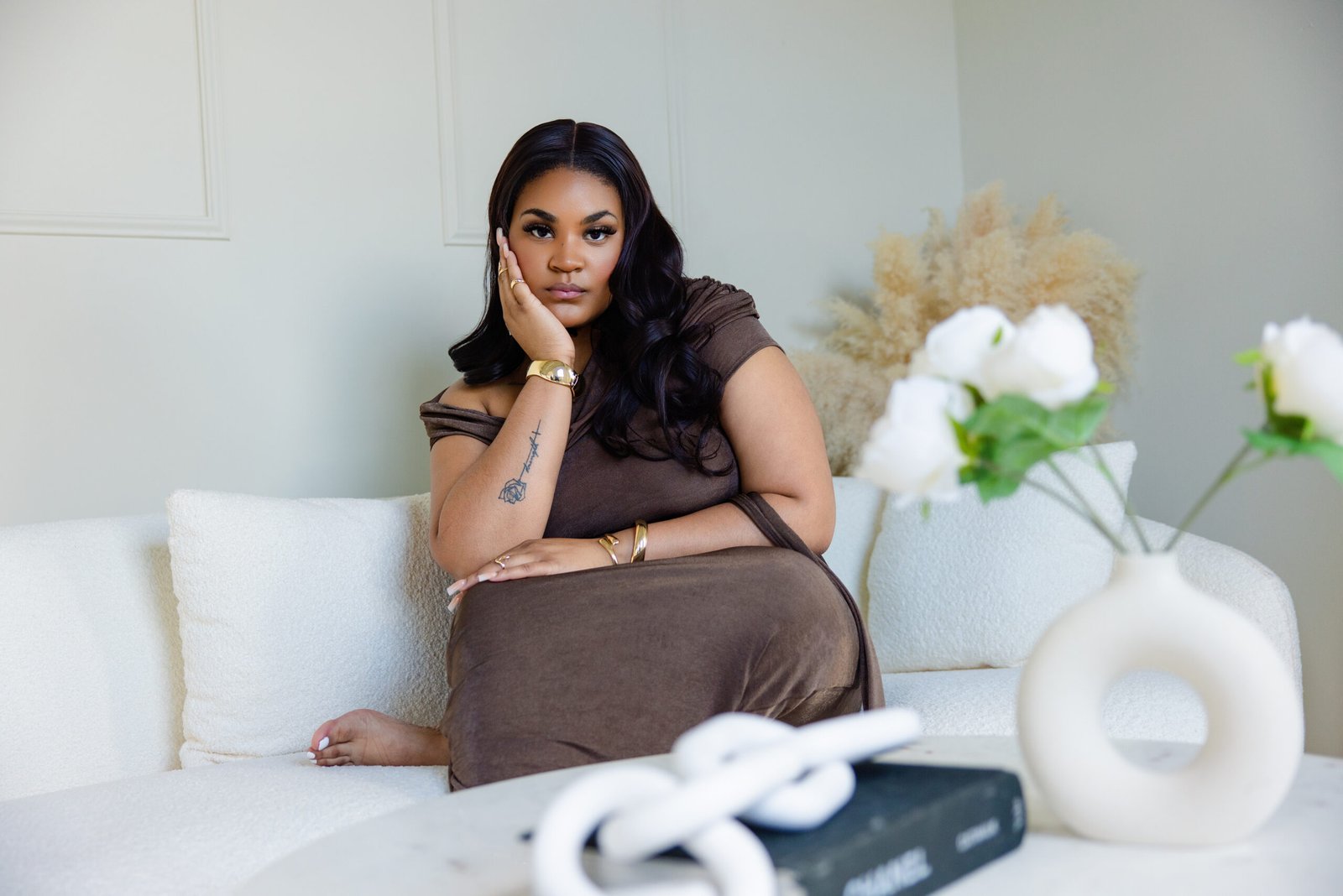 Personal branding portrait of a model in a brown dress seated on a white sofa, resting her chin in her hand