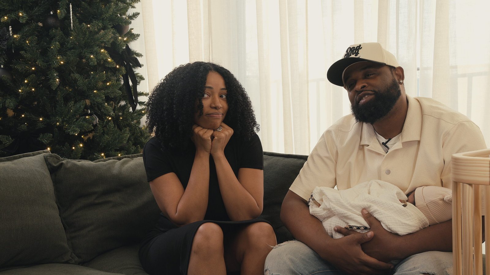 Couple sharing a lighthearted moment on the couch with their newborn beside a Christmas tree during a family documentary interview