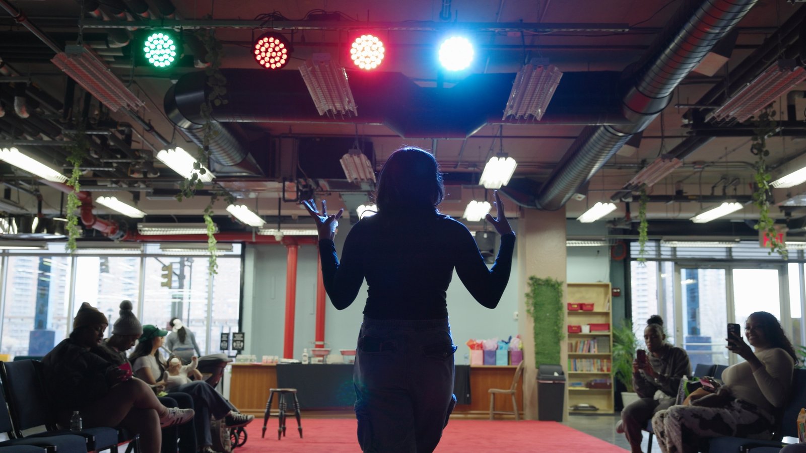 Chelsea of The Christian Doula Experience addressing mothers at her Mommy and Me event, filmed from behind under colorful stage lights
