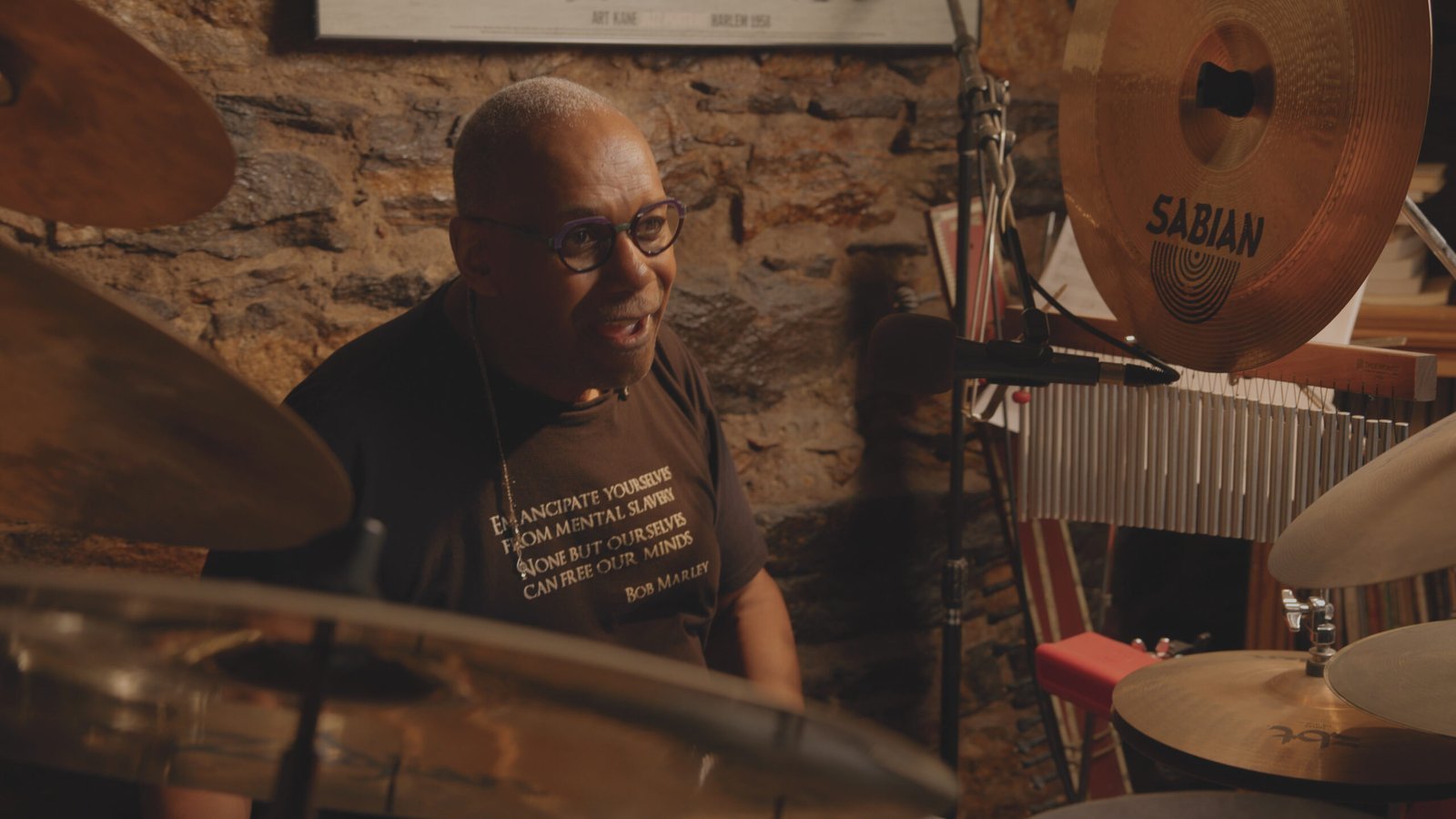 Jazz drummer speaking from behind his drum kit during a documentary interview in a stone-walled music studio